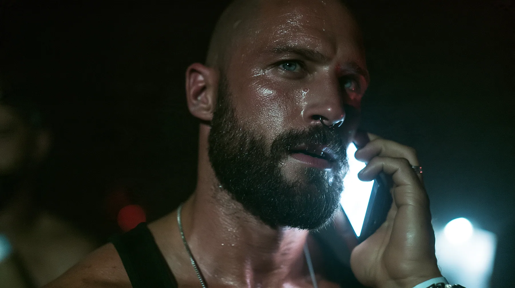 extreme close-up of a phone pressed to the ear of a broad-shouldered Caucasian man in his early thirties with a shaved head, a thick short dark beard, a square jaw, heavy brows and pale blue eyes, the build of a man…