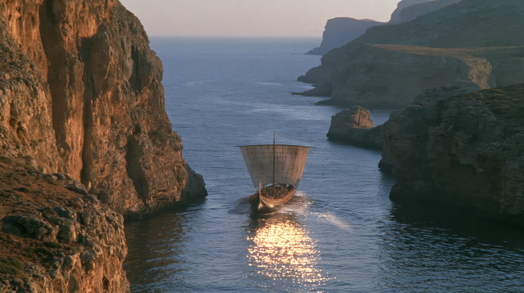 aerial of a long black ancient Greek galley with a single mast, a square sail and two banks of oars leaving a narrow strait of cliffs for the open sea, sunrise, warm gold light and long shadows, spray lit gold,…