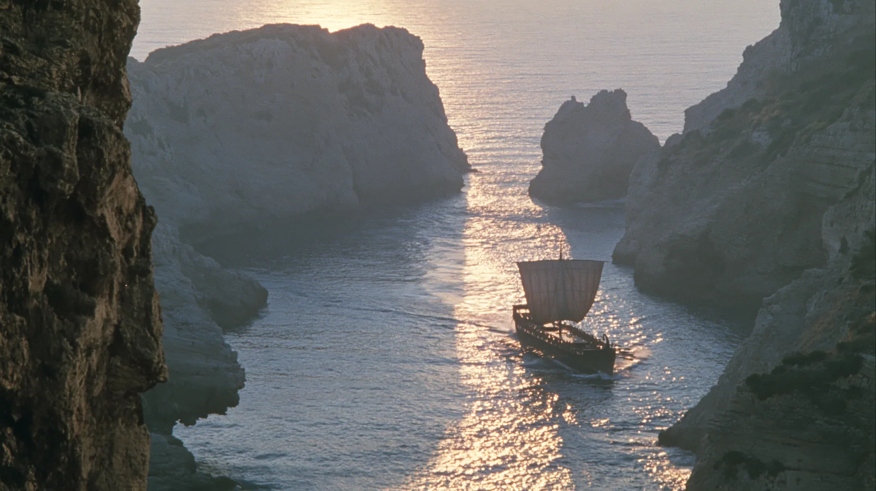 aerial of a long black ancient Greek galley with a single mast, a square sail and two banks of oars leaving a narrow strait of cliffs for the open sea, sunrise, warm gold light and long shadows, spray lit gold,…