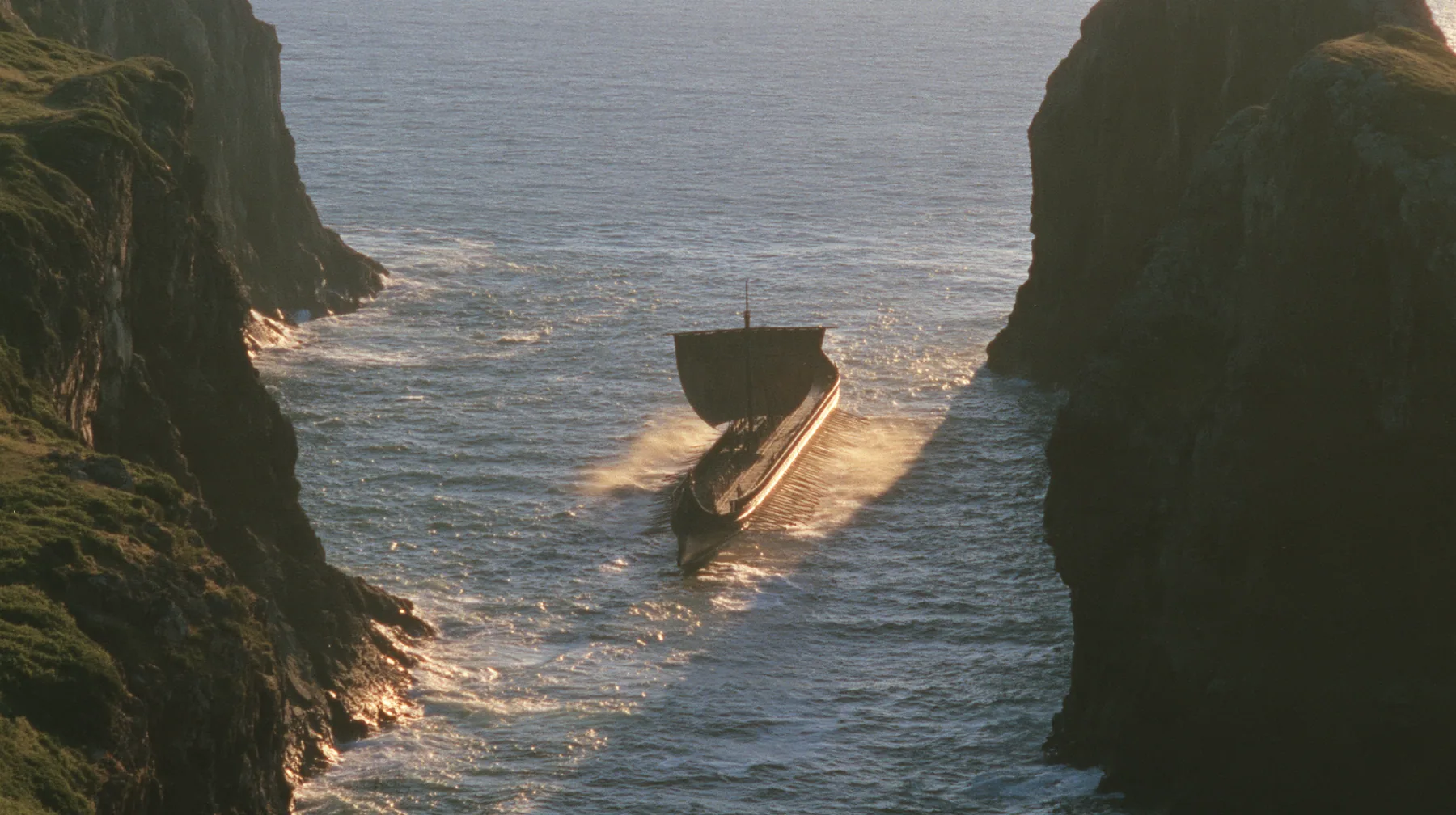 aerial of a long black ancient Greek galley with a single mast, a square sail and two banks of oars leaving a narrow strait of cliffs for the open sea, sunrise, warm gold light and long shadows, spray lit gold,…