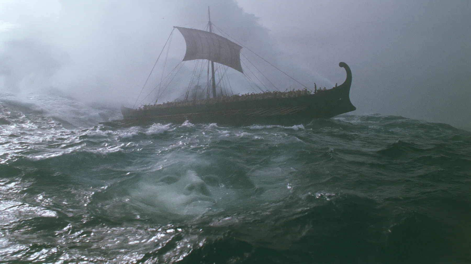 wide shot of a long black ancient Greek galley with a single mast, a square sail and two banks of oars lifted high on a rising mountain of water, and through the translucent face of the swell a colossal pale woman's…