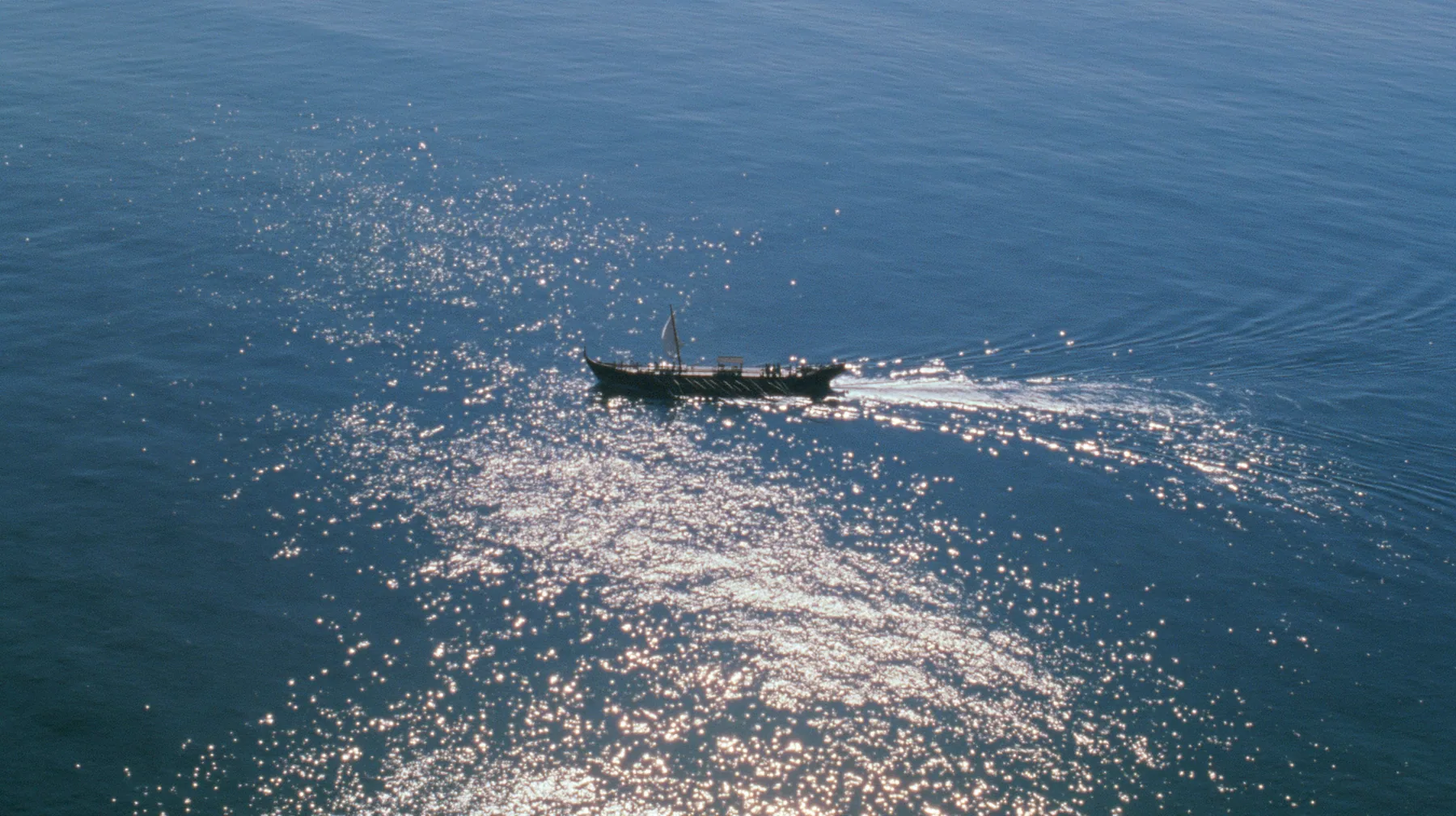extreme high aerial of a long black ancient Greek galley with a single mast, a square sail and two banks of oars as a tiny dark shape on a vast sparkling morning sea, its wake a thin white line, clear morning light…