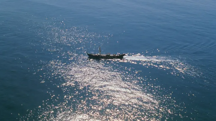 extreme high aerial of a long black ancient Greek galley with a single mast, a square sail and two banks of oars as a tiny dark shape on a vast sparkling morning sea, its wake a thin white line, clear morning light…