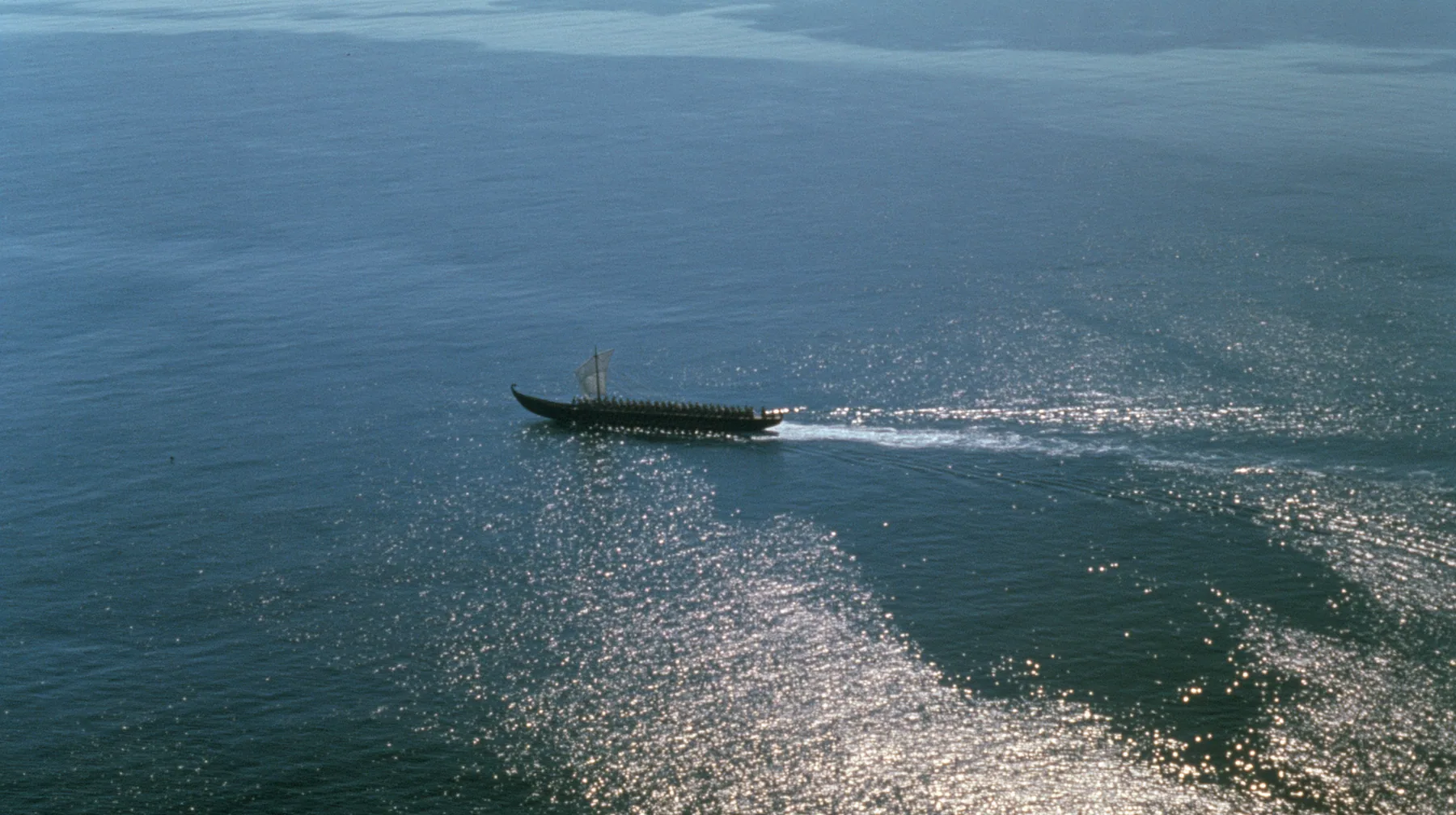 extreme high aerial of a long black ancient Greek galley with a single mast, a square sail and two banks of oars as a tiny dark shape on a vast sparkling morning sea, its wake a thin white line, clear morning light…