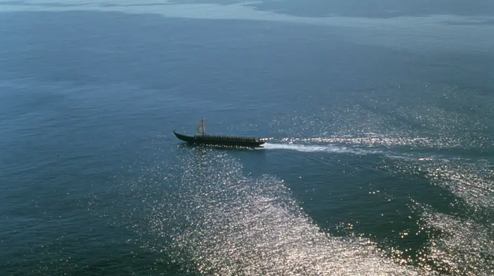 extreme high aerial of a long black ancient Greek galley with a single mast, a square sail and two banks of oars as a tiny dark shape on a vast sparkling morning sea, its wake a thin white line, clear morning light…