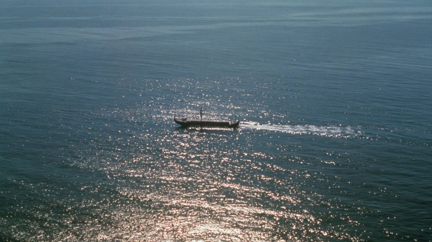 extreme high aerial of a long black ancient Greek galley with a single mast, a square sail and two banks of oars as a tiny dark shape on a vast sparkling morning sea, its wake a thin white line, clear morning light…
