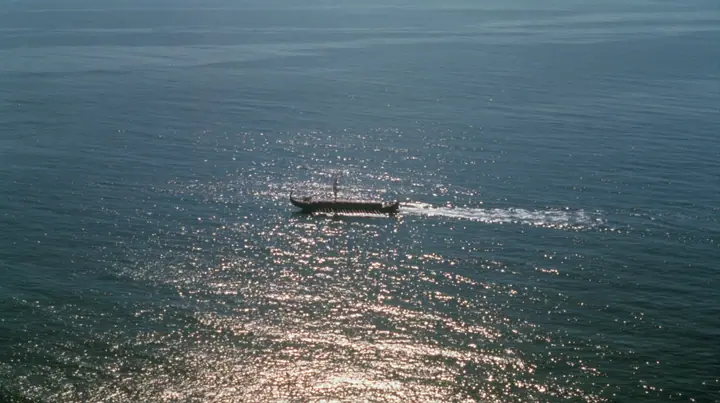 extreme high aerial of a long black ancient Greek galley with a single mast, a square sail and two banks of oars as a tiny dark shape on a vast sparkling morning sea, its wake a thin white line, clear morning light…