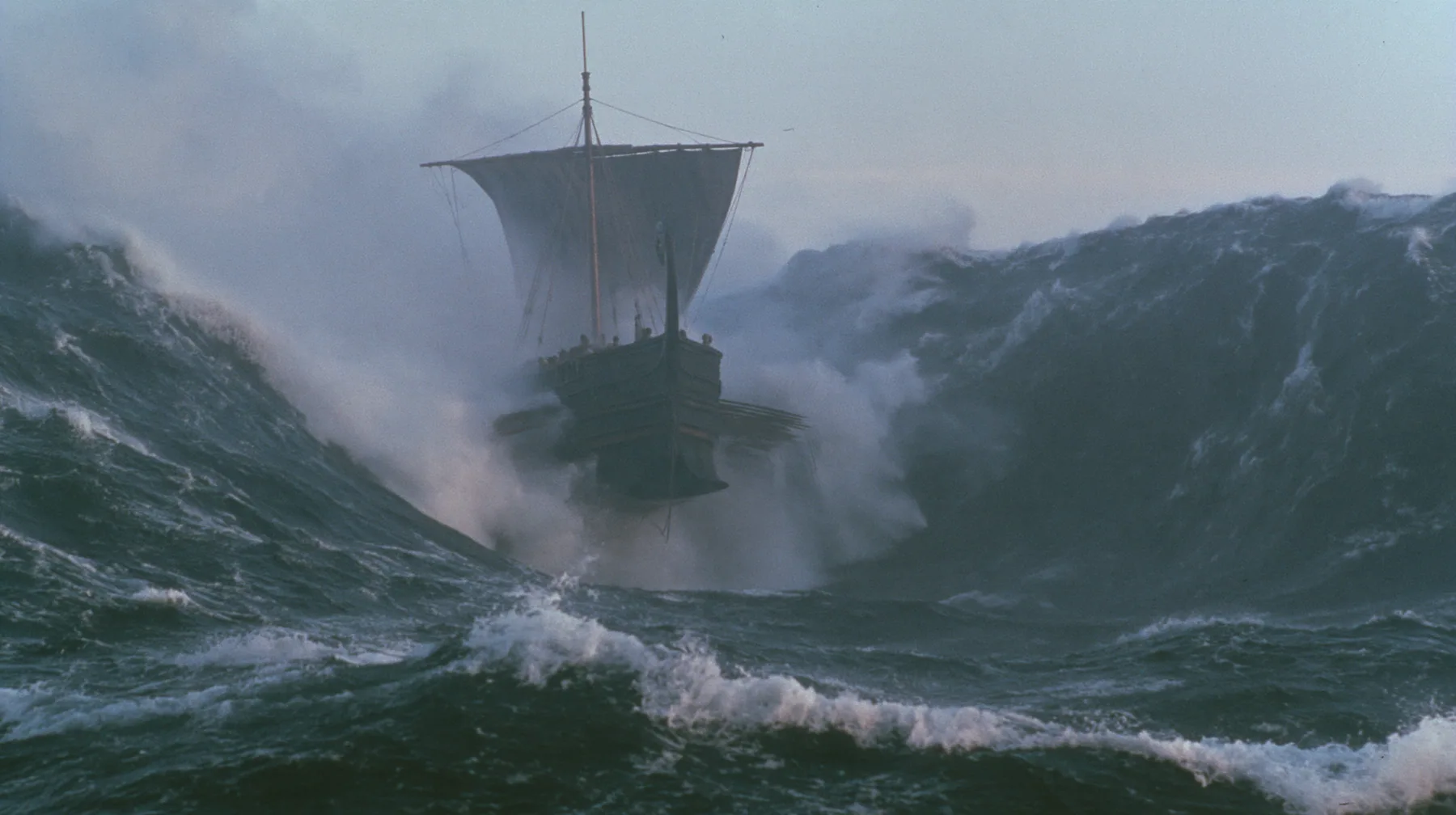 wide shot of a long black ancient Greek galley with a single mast, a square sail and two banks of oars dropping into an enormous trough as the sea falls away around it, walls of dark water on both sides, grey first…