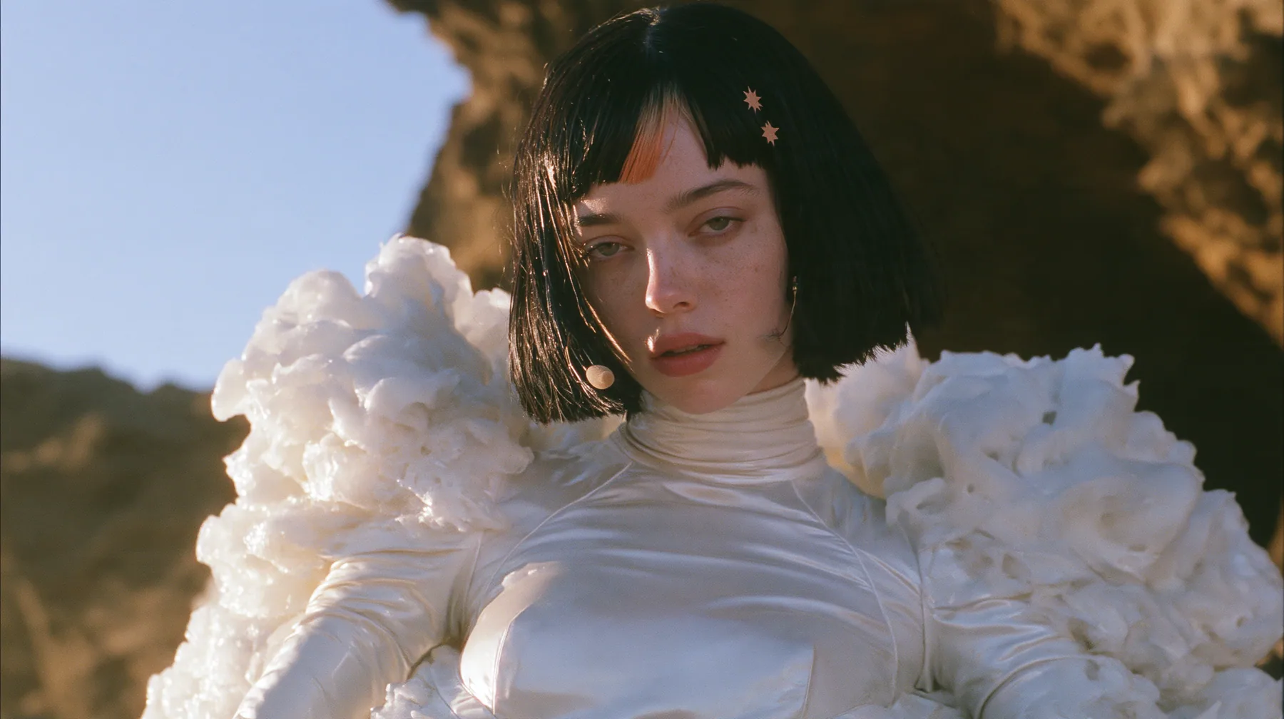 medium shot of a Caucasian American woman in her mid-twenties with pale skin and light freckles, a glossy black blunt jaw-length bob with heavy straight bangs and one clay-orange streak through the bangs, a small…