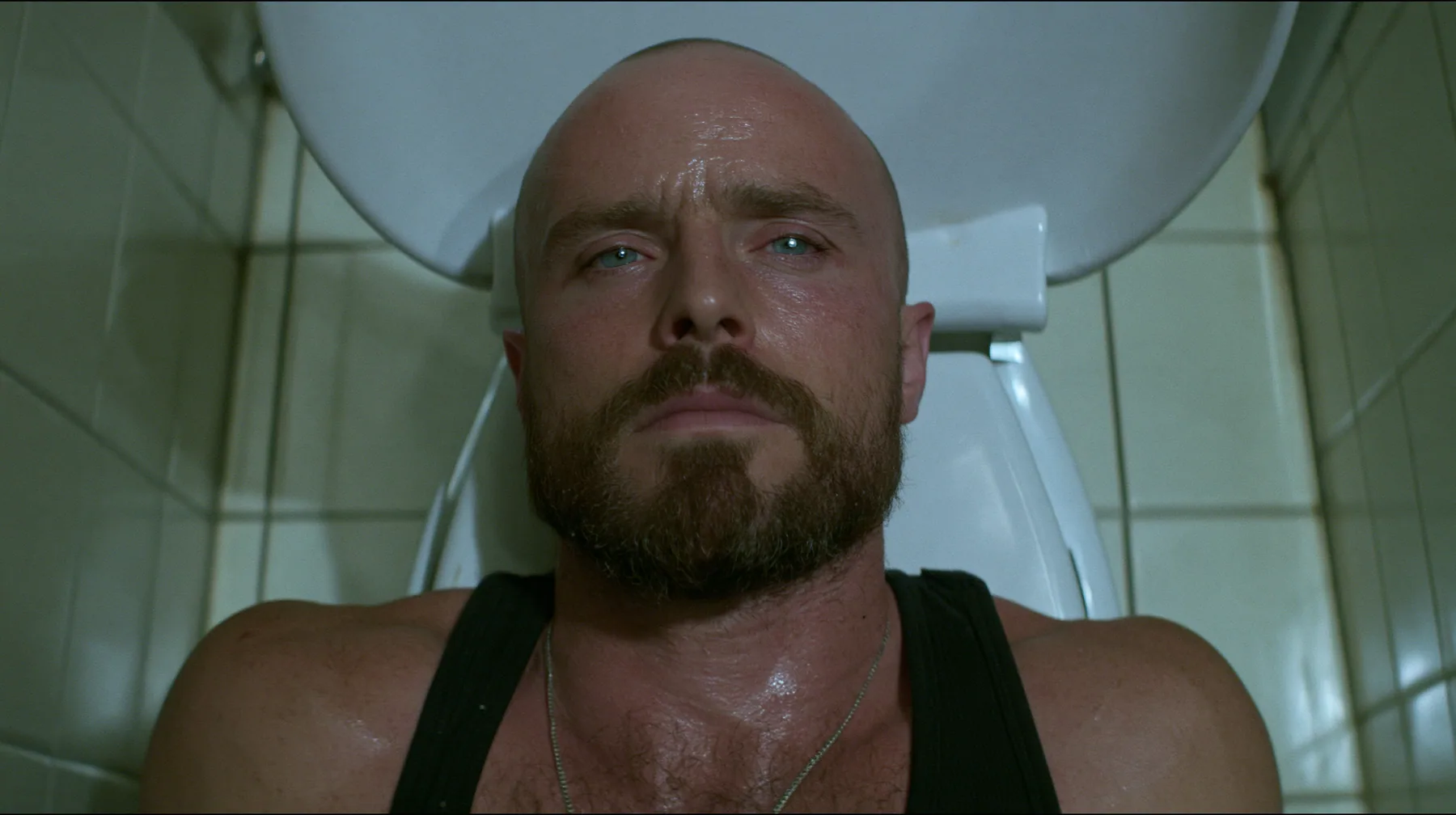 straight-down shot of a broad-shouldered Caucasian man in his early thirties with a shaved head, a thick short dark beard, a square jaw, heavy brows and pale blue eyes, the build of a man who lifts, in a black tank…