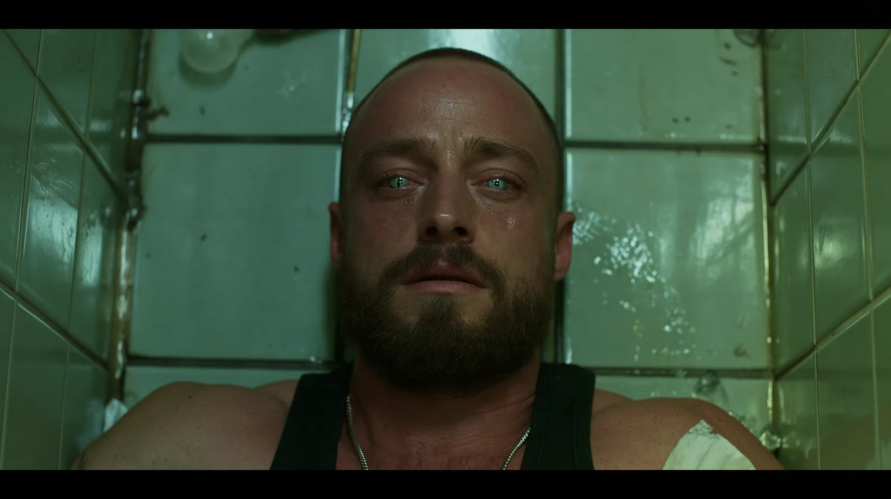 straight-down shot of a broad-shouldered Caucasian man in his early thirties with a shaved head, a thick short dark beard, a square jaw, heavy brows and pale blue eyes, the build of a man who lifts, in a black tank…