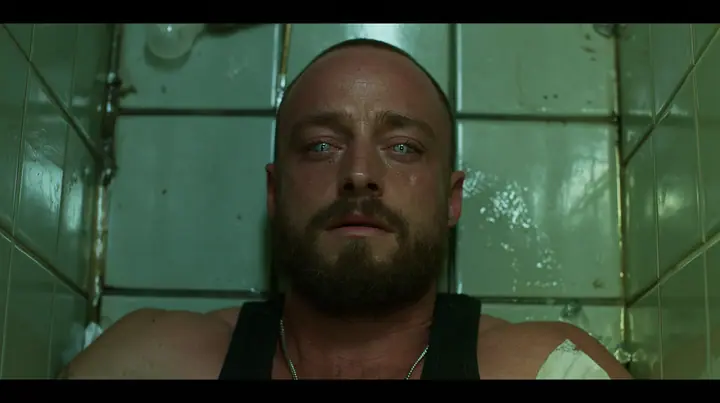 straight-down shot of a broad-shouldered Caucasian man in his early thirties with a shaved head, a thick short dark beard, a square jaw, heavy brows and pale blue eyes, the build of a man who lifts, in a black tank…