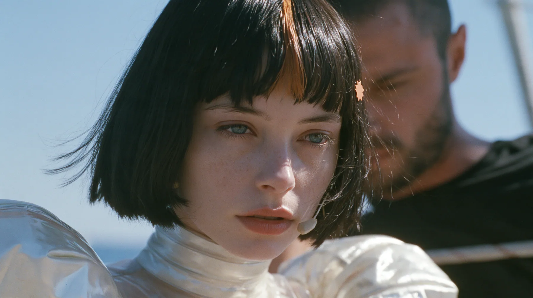 close-up of a Caucasian American woman in her mid-twenties with pale skin and light freckles, a glossy black blunt jaw-length bob with heavy straight bangs and one clay-orange streak through the bangs, a small flat…