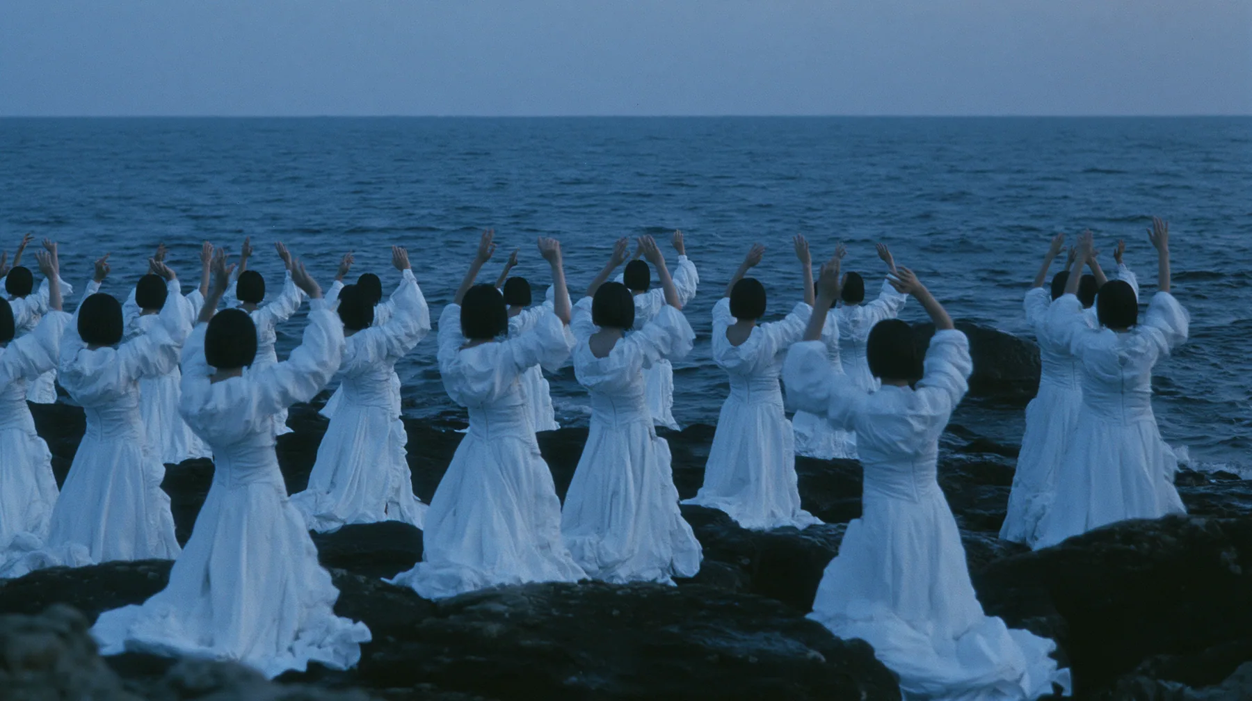 wide shot of hundreds of identical bare-headed women with glossy black bobs in structured white couture gowns on black sea rocks, all raising their arms toward a passing black galley, open ancient sea to the horizon…