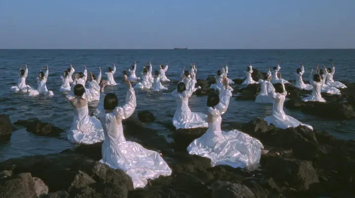 wide shot of hundreds of identical bare-headed women with glossy black bobs in structured white couture gowns on black sea rocks, all raising their arms toward a passing black galley, open ancient sea to the horizon…