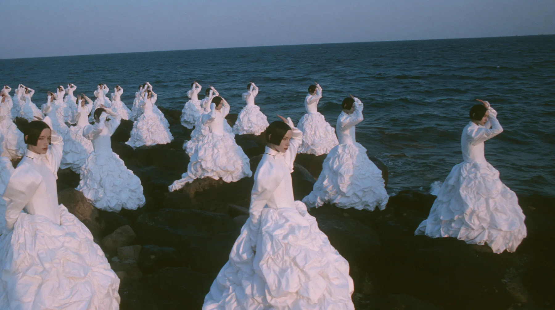 wide shot of hundreds of identical bare-headed women with glossy black bobs in structured white couture gowns on black sea rocks, all raising their arms toward a passing black galley, open ancient sea to the horizon…