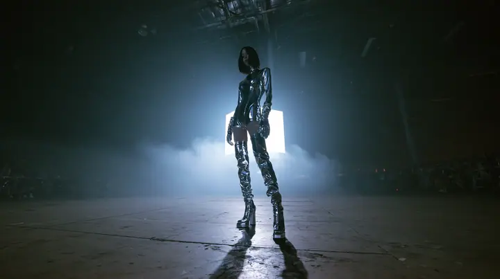 extreme wide shot of a vast dark warehouse rave, and alone in the middle of the empty concrete floor a young Caucasian American woman with pale skin, a glossy black blunt jaw-length bob with heavy straight bangs and…