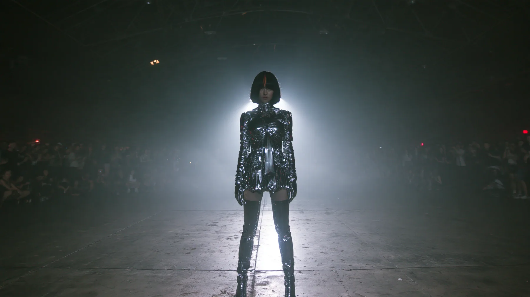 extreme wide shot of a vast dark warehouse rave, and alone in the middle of the empty concrete floor a young Caucasian American woman with pale skin, a glossy black blunt jaw-length bob with heavy straight bangs and…