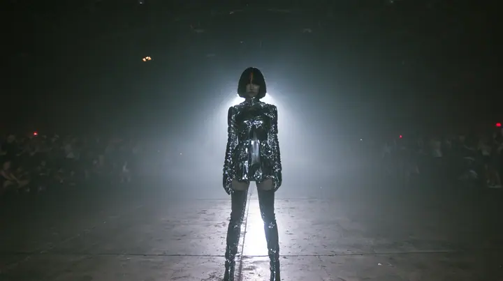 extreme wide shot of a vast dark warehouse rave, and alone in the middle of the empty concrete floor a young Caucasian American woman with pale skin, a glossy black blunt jaw-length bob with heavy straight bangs and…