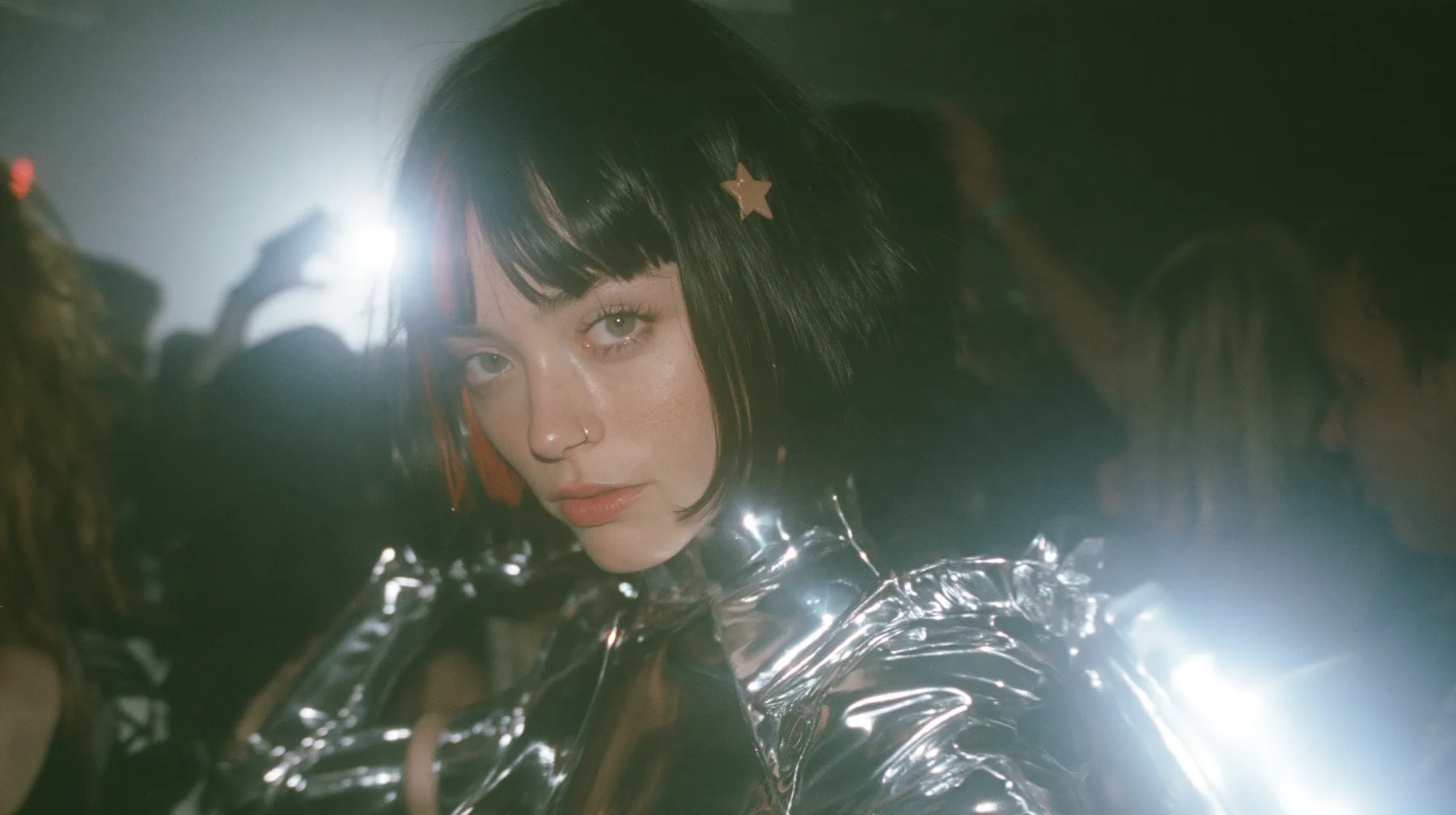 cinematic 35mm film still, wide shot at eye level: a warehouse rave in fog, the dancers parted into a circle, and in the circle stands a young Caucasian American woman with pale skin, a glossy black blunt jaw-length…
