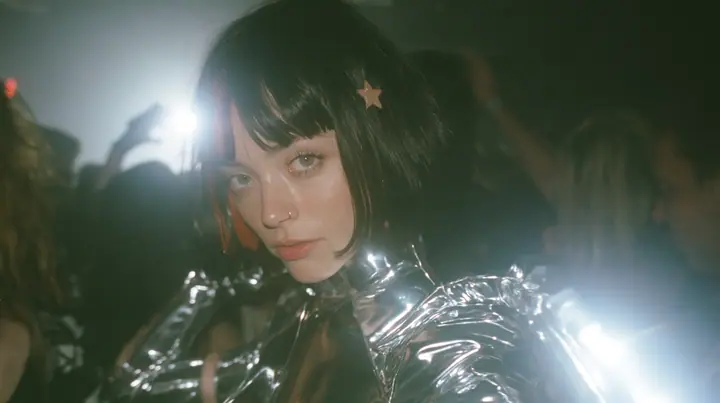 cinematic 35mm film still, wide shot at eye level: a warehouse rave in fog, the dancers parted into a circle, and in the circle stands a young Caucasian American woman with pale skin, a glossy black blunt jaw-length…