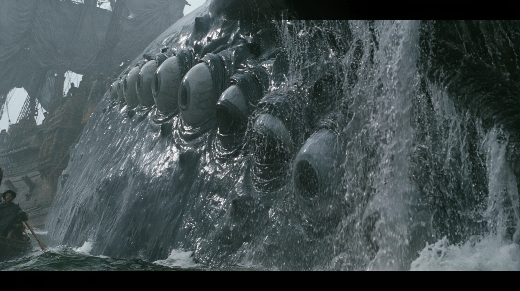 close shot along the flank of a gigantic sea creature breaching beside a wooden galley, its glistening dark skin covered in rows of enormous wet eyes with pale irises, the eyes opening one after another, water…