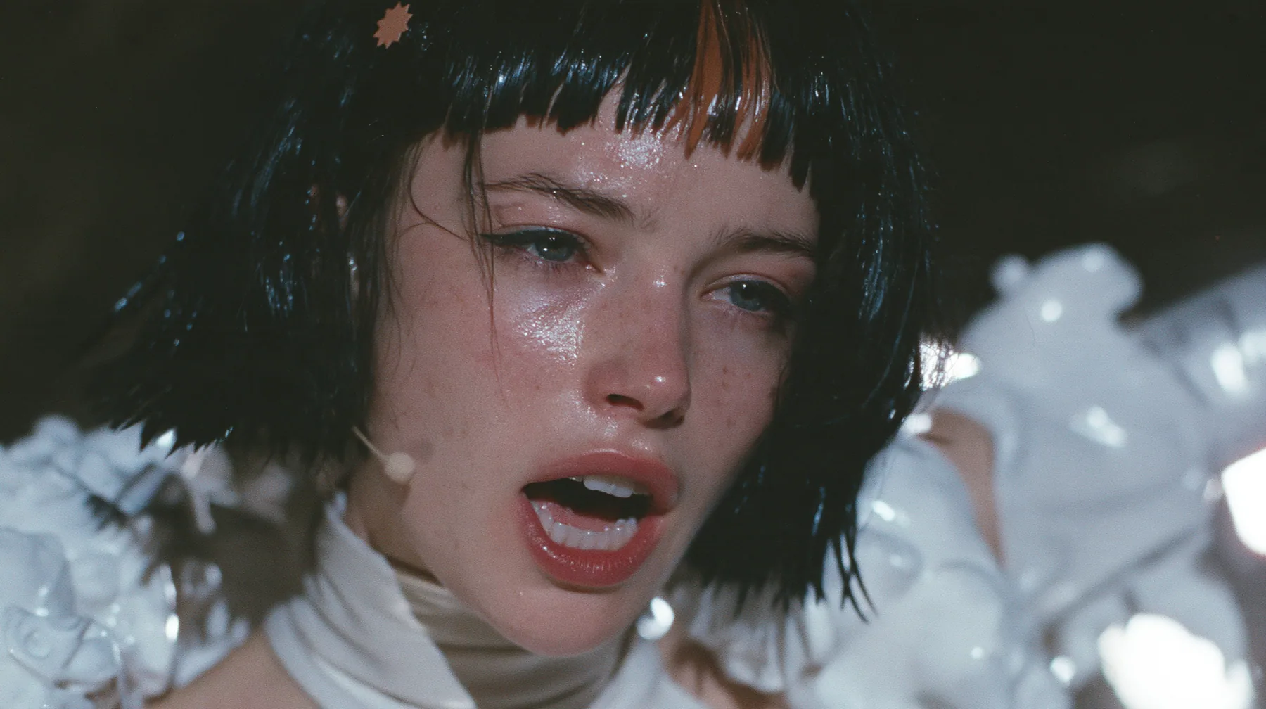 medium close-up of a Caucasian American woman in her mid-twenties with pale skin and light freckles, a glossy black blunt jaw-length bob with heavy straight bangs and one clay-orange streak through the bangs, a small…