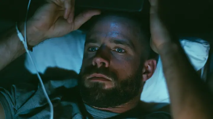 close-up of a broad-shouldered Caucasian man in his early thirties with a shaved head, a thick short dark beard, a square jaw, heavy brows and pale blue eyes, the build of a man who lifts, in a grey t-shirt, lying…