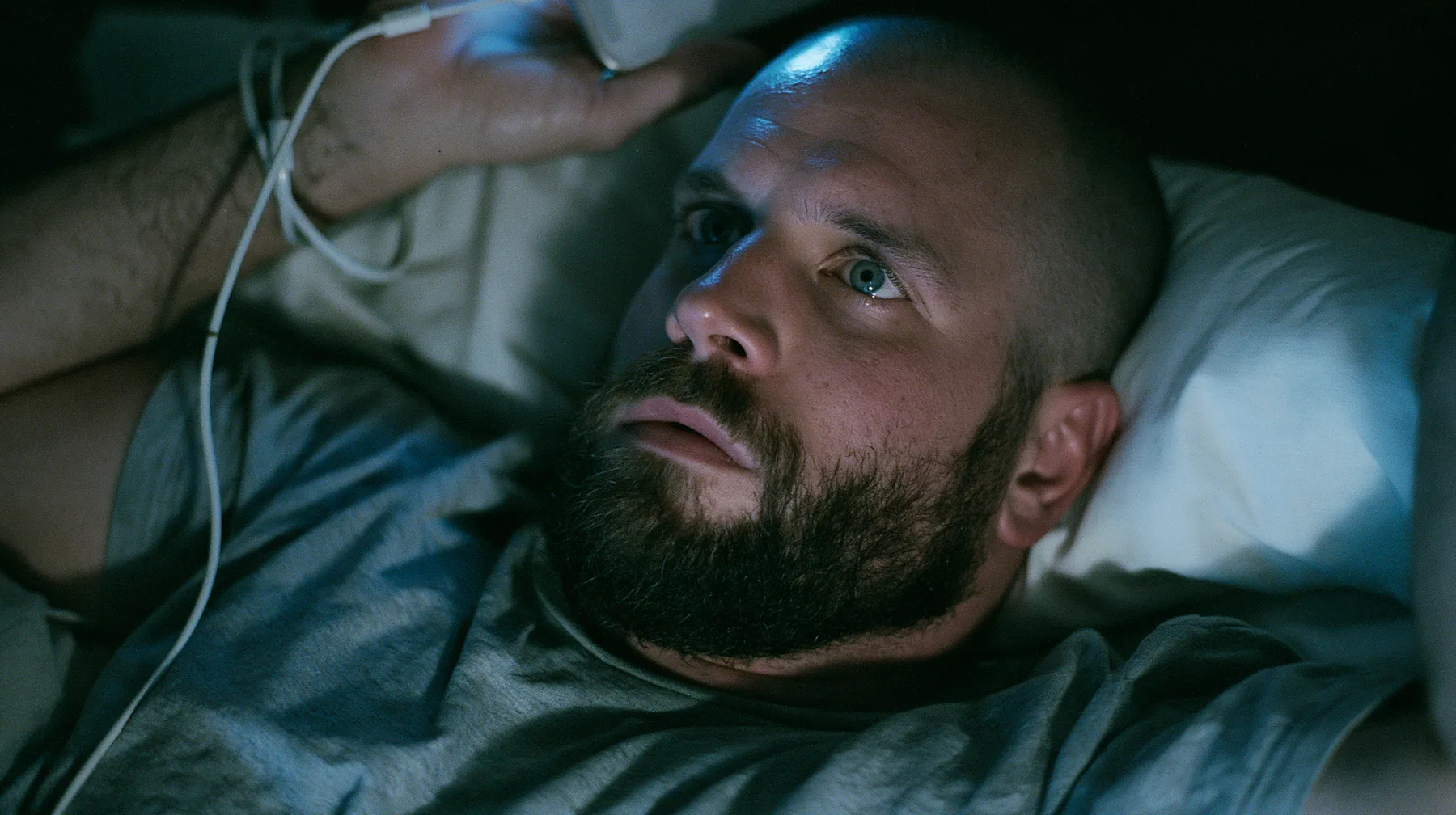 close-up of a broad-shouldered Caucasian man in his early thirties with a shaved head, a thick short dark beard, a square jaw, heavy brows and pale blue eyes, the build of a man who lifts, in a grey t-shirt, lying…