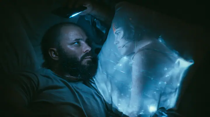top-down shot of a broad-shouldered Caucasian man in his early thirties with a shaved head, a thick short dark beard, a square jaw, heavy brows and pale blue eyes, the build of a man who lifts, in a grey t-shirt,…
