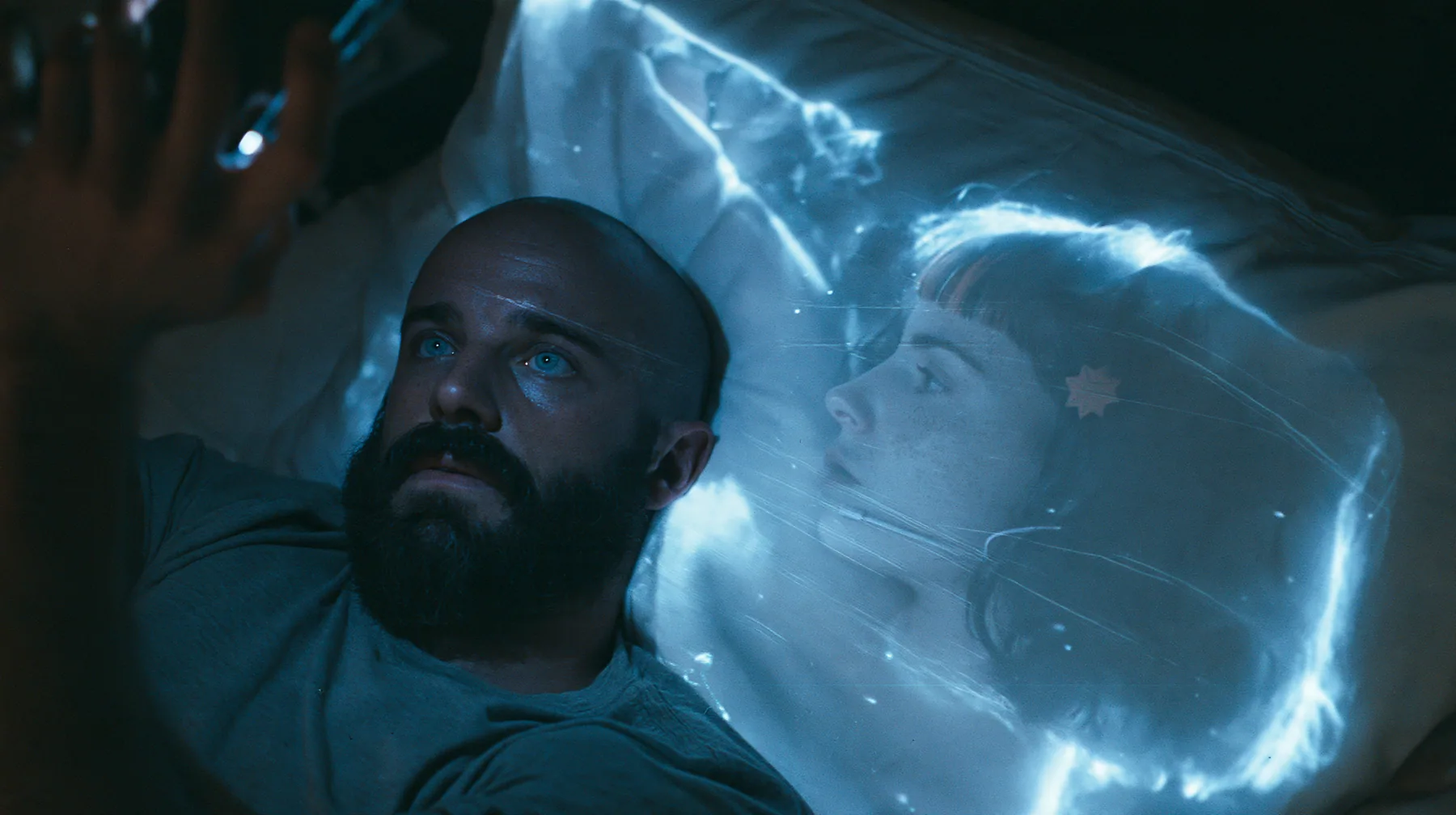 top-down shot of a broad-shouldered Caucasian man in his early thirties with a shaved head, a thick short dark beard, a square jaw, heavy brows and pale blue eyes, the build of a man who lifts, in a grey t-shirt,…