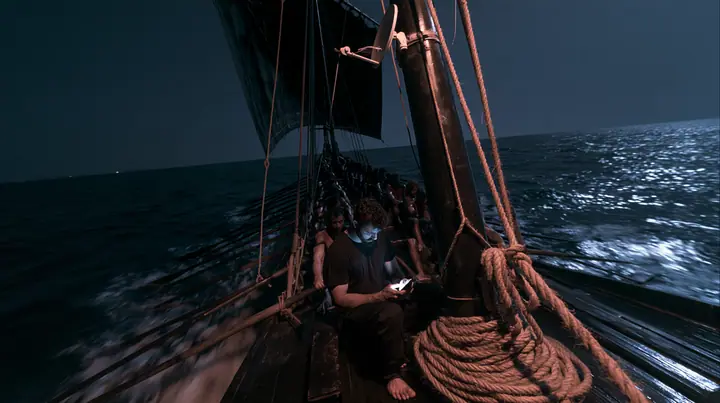 wide shot from the stern along the deck of a long black ancient Greek galley with a single mast, a square sail and two banks of oars at night, thirty rowers on two long rows of benches pulling their oars, a tall mast…