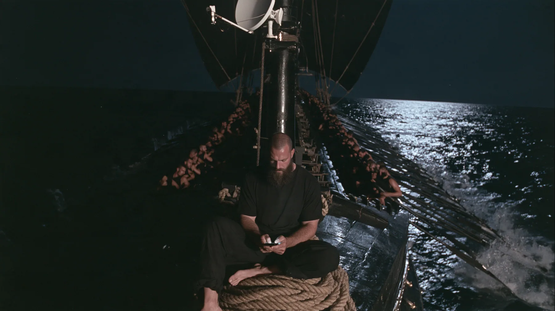 wide shot from the stern along the deck of a long black ancient Greek galley with a single mast, a square sail and two banks of oars at night, thirty rowers on two long rows of benches pulling their oars, a tall mast…