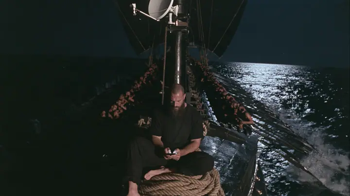 wide shot from the stern along the deck of a long black ancient Greek galley with a single mast, a square sail and two banks of oars at night, thirty rowers on two long rows of benches pulling their oars, a tall mast…