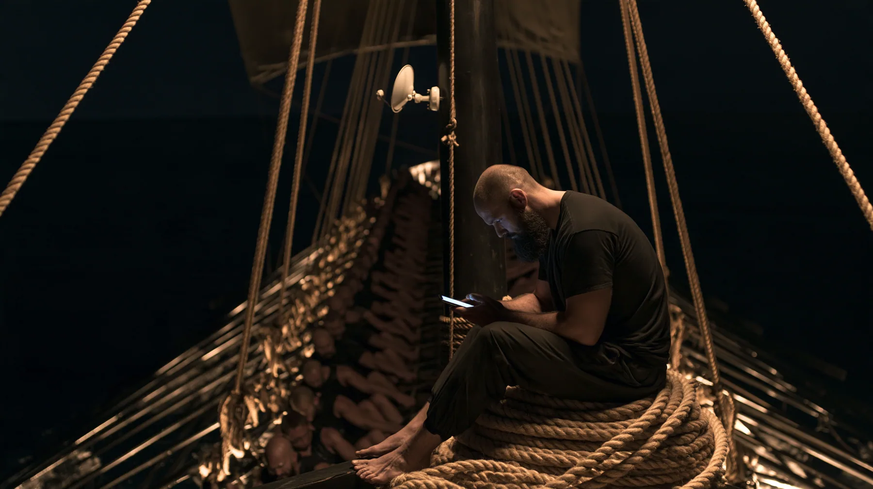 wide shot from the stern along the deck of a long black ancient Greek galley with a single mast, a square sail and two banks of oars at night, thirty rowers on two long rows of benches pulling their oars, a tall mast…