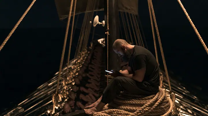 wide shot from the stern along the deck of a long black ancient Greek galley with a single mast, a square sail and two banks of oars at night, thirty rowers on two long rows of benches pulling their oars, a tall mast…