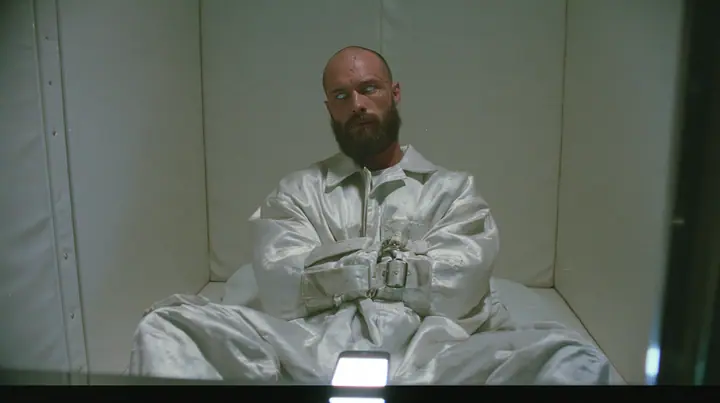 a broad-shouldered Caucasian man in his early thirties with a shaved head, a thick short dark beard, a square jaw, heavy brows and pale blue eyes, the build of a man who lifts, in a white canvas straitjacket, its…