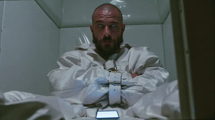 a broad-shouldered Caucasian man in his early thirties with a shaved head, a thick short dark beard, a square jaw, heavy brows and pale blue eyes, the build of a man who lifts, in a white canvas straitjacket, its…