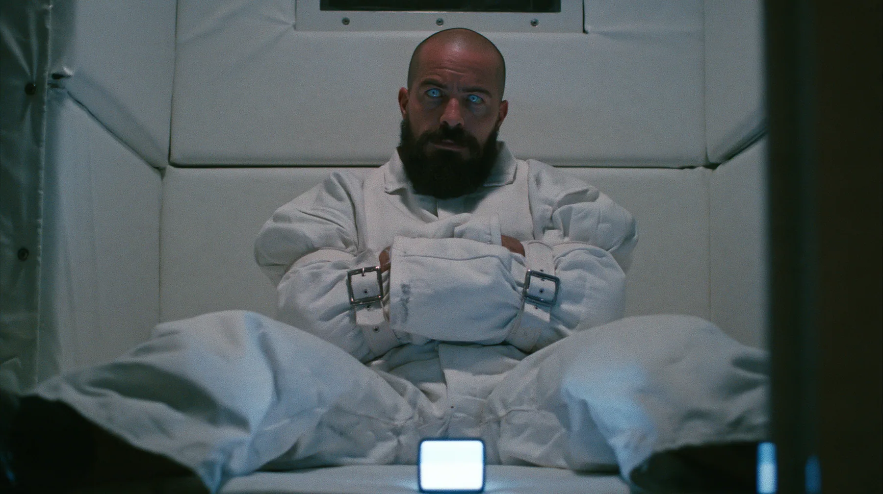 a broad-shouldered Caucasian man in his early thirties with a shaved head, a thick short dark beard, a square jaw, heavy brows and pale blue eyes, the build of a man who lifts, in a white canvas straitjacket, its…