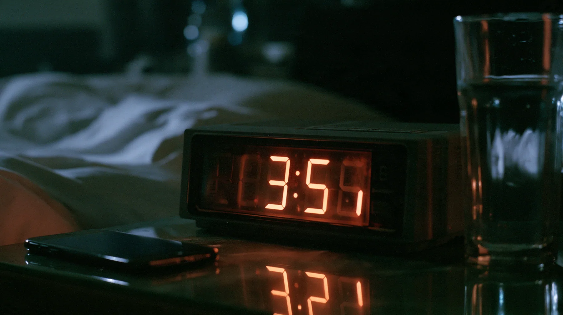 macro of a bedside digital alarm clock in a dark bedroom, its big seven-segment display glowing orange, a phone lying face up beside it lighting the ceiling, a glass of water, shallow depth of field, a dark bedroom…