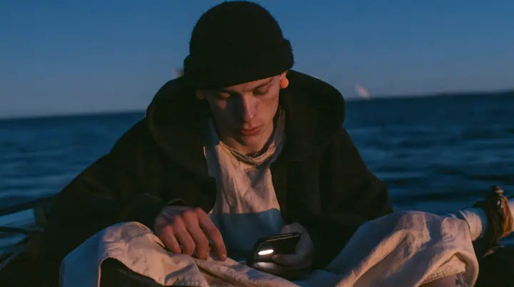 medium close-up of a gaunt pale young rower with dark circles under his eyes in a black knit beanie and a black hoodie over a linen tunic, a small orange foam earplug in each ear, sitting on a rowing bench of an…