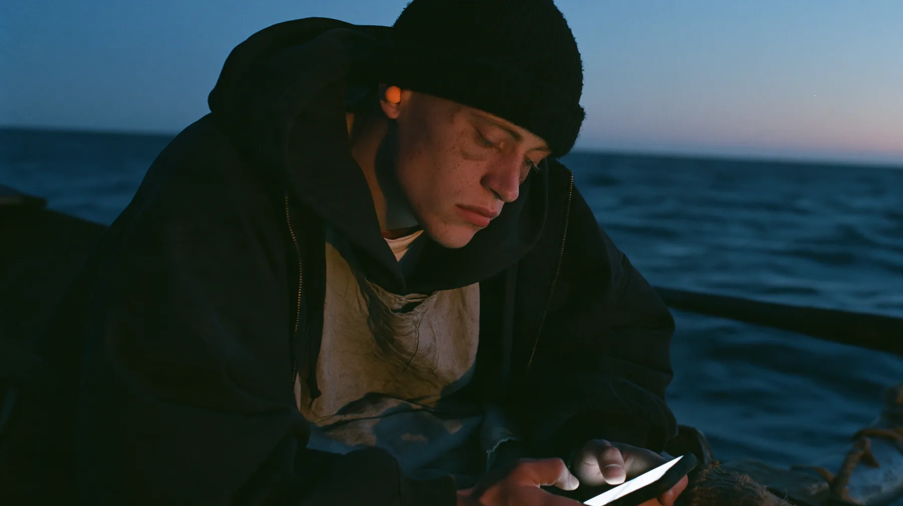 medium close-up of a gaunt pale young rower with dark circles under his eyes in a black knit beanie and a black hoodie over a linen tunic, a small orange foam earplug in each ear, sitting on a rowing bench of an…