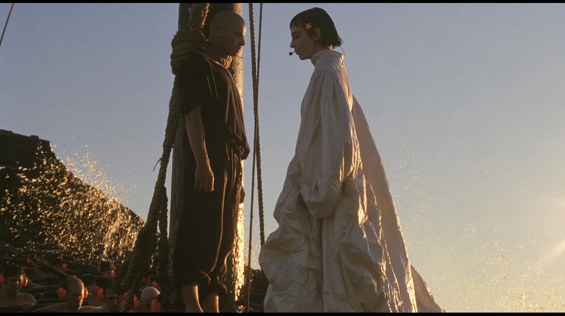 side view at sunrise on the deck of a long black ancient Greek galley with a single mast, a square sail and two banks of oars: a broad-shouldered Caucasian man in his early thirties with a shaved head, a thick short…