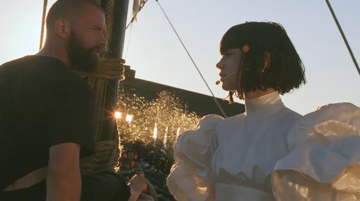 side view at sunrise on the deck of a long black ancient Greek galley with a single mast, a square sail and two banks of oars: a broad-shouldered Caucasian man in his early thirties with a shaved head, a thick short…