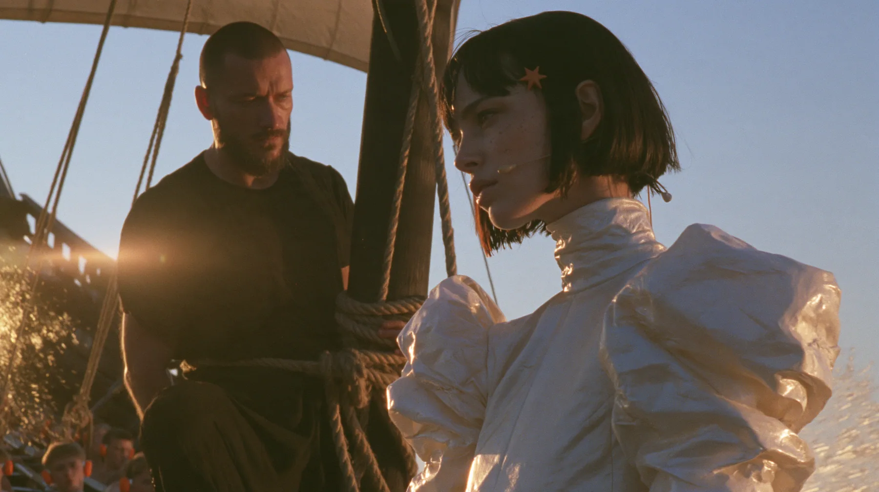 side view at sunrise on the deck of a long black ancient Greek galley with a single mast, a square sail and two banks of oars: a broad-shouldered Caucasian man in his early thirties with a shaved head, a thick short…