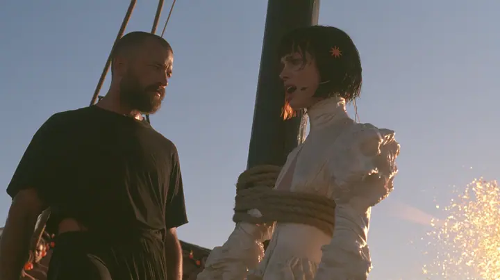 side view at sunrise on the deck of a long black ancient Greek galley with a single mast, a square sail and two banks of oars: a Caucasian American woman in her mid-twenties with pale skin and light freckles, a…