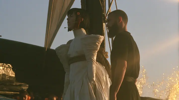 side view at sunrise on the deck of a long black ancient Greek galley with a single mast, a square sail and two banks of oars: a Caucasian American woman in her mid-twenties with pale skin and light freckles, a…
