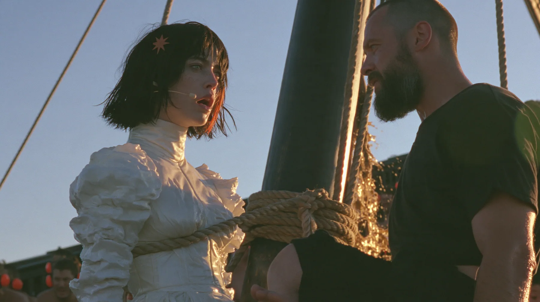 side view at sunrise on the deck of a long black ancient Greek galley with a single mast, a square sail and two banks of oars: a Caucasian American woman in her mid-twenties with pale skin and light freckles, a…
