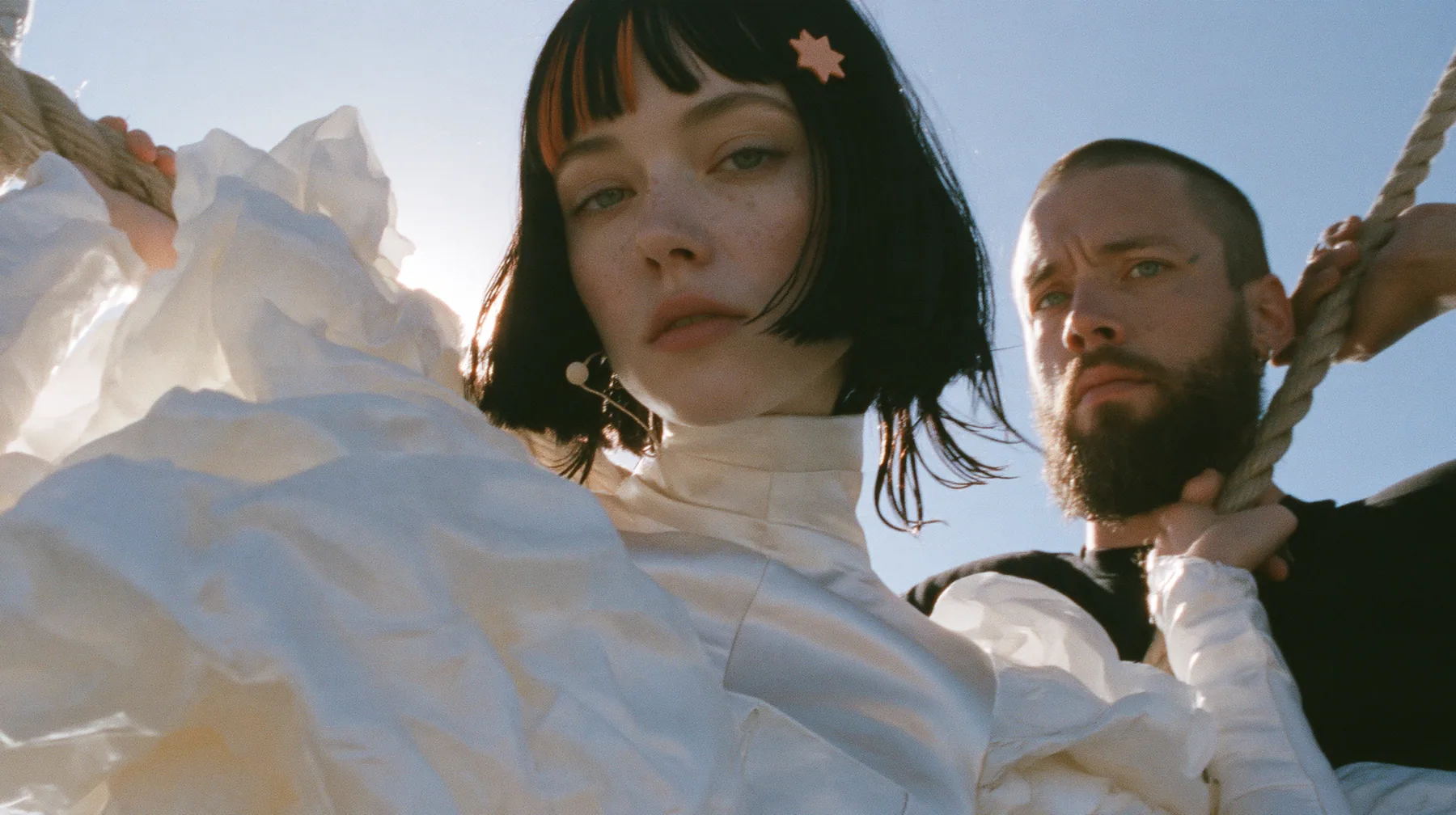 low angle of a Caucasian American woman in her mid-twenties with pale skin and light freckles, a glossy black blunt jaw-length bob with heavy straight bangs and one clay-orange streak through the bangs, a small flat…