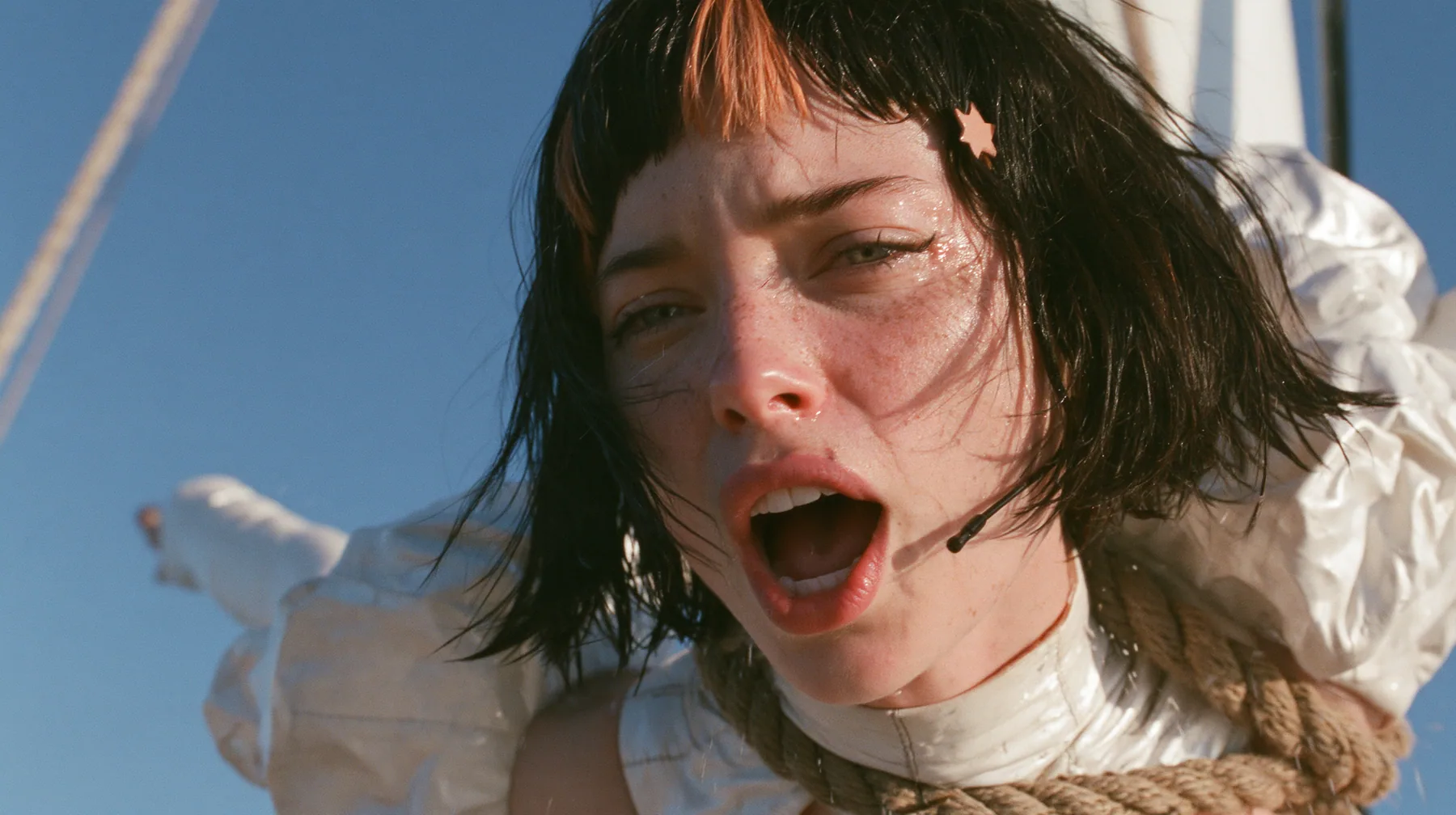 close-up of a Caucasian American woman in her mid-twenties with pale skin and light freckles, a glossy black blunt jaw-length bob with heavy straight bangs and one clay-orange streak through the bangs, a small flat…